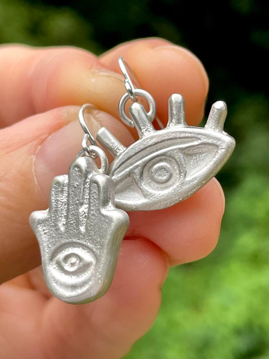 Silver Hamsa hand and eye earrings held between fingers with a blurred green background