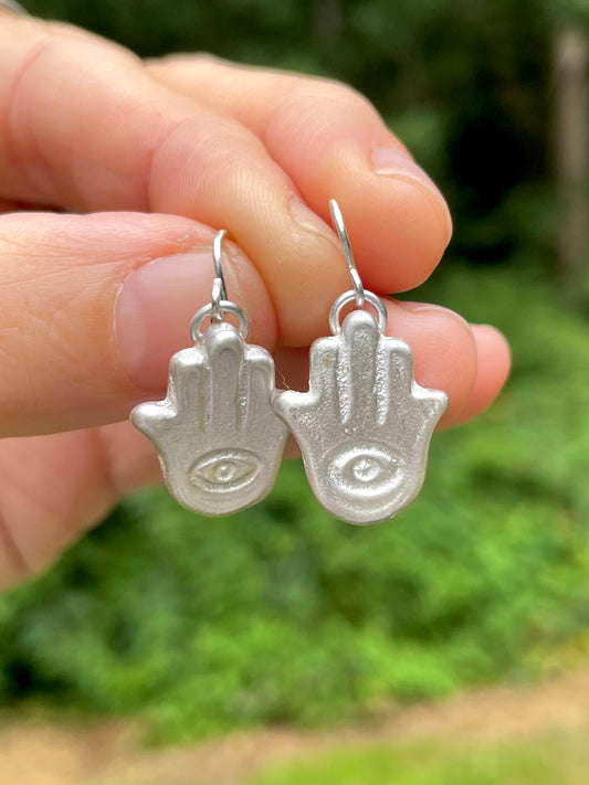 Silver sandcast Hamsa hand earrings held between fingers with a blurred green background