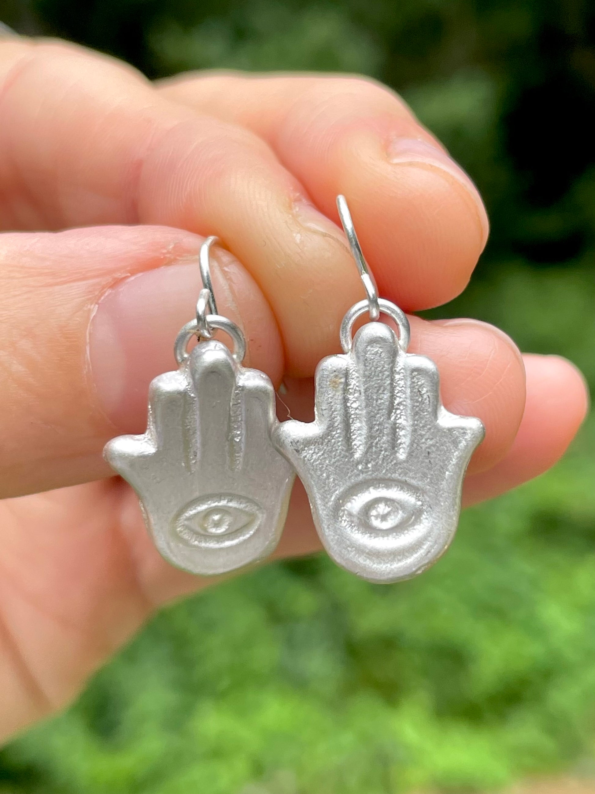 Silver sandcast Hamsa hand earrings held between fingers with a blurred green background