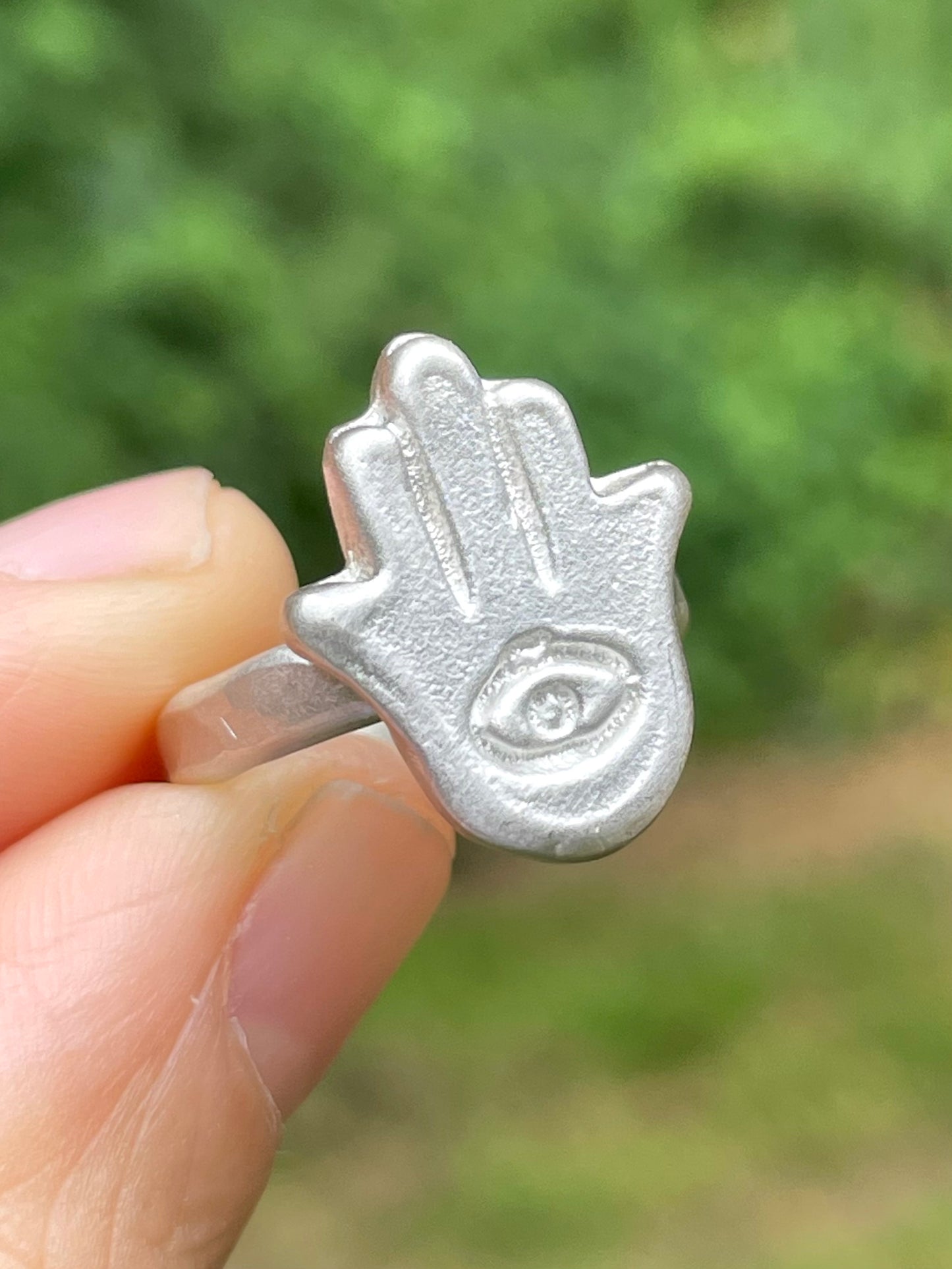 Silver sandcast Hamsa hand ring held between fingers with a blurred green background
