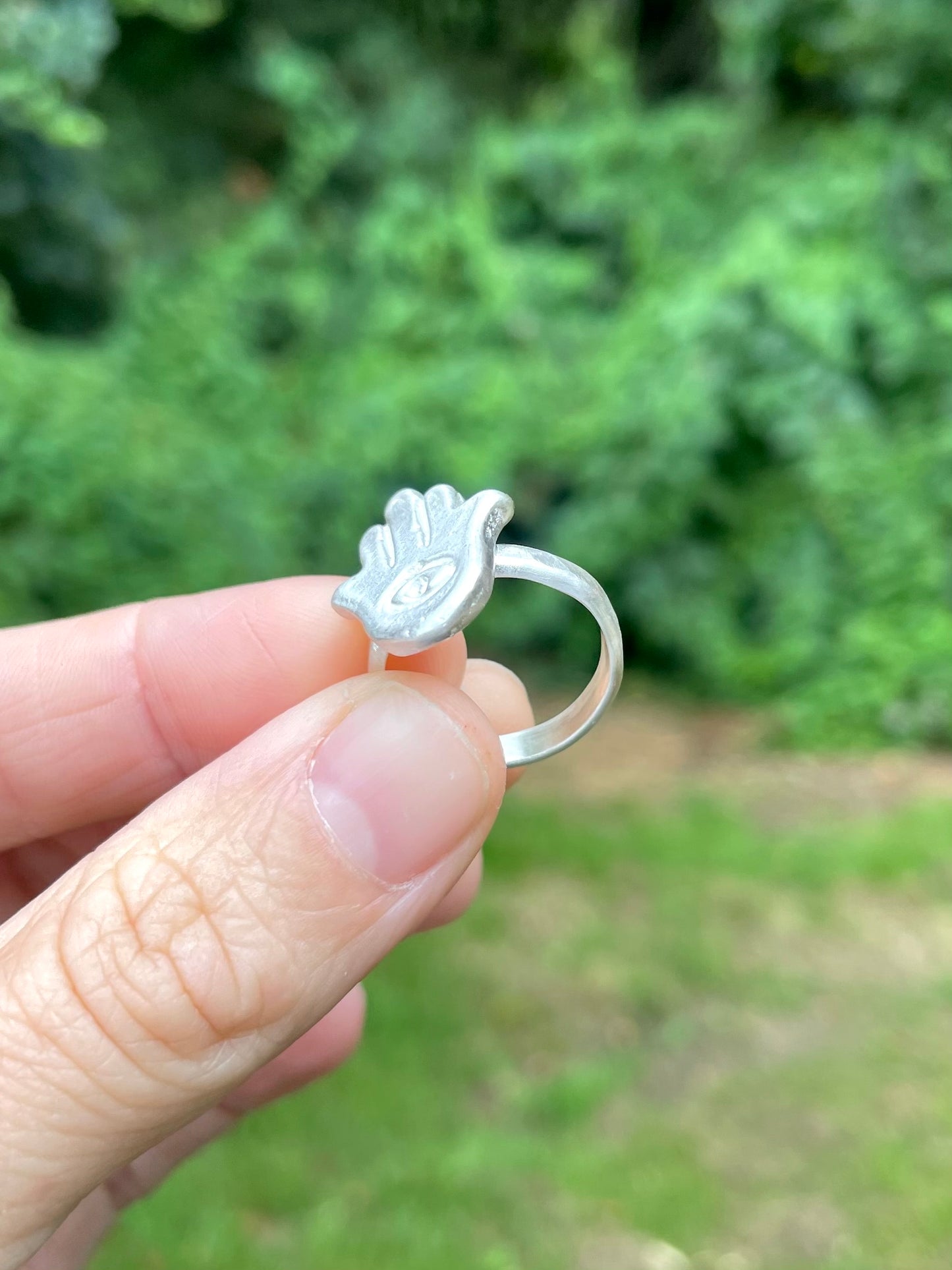 Silver sandcast hamsa hand ring held by a hand against a blurred green outdoor background