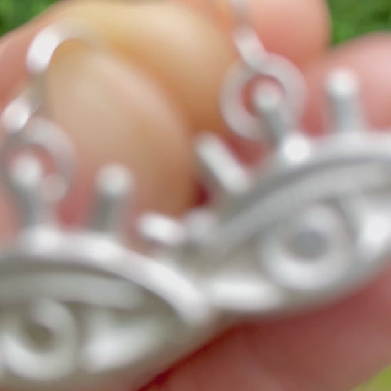 silver sandcast eye shaped earrings held in a hand with a blurred green grass background
