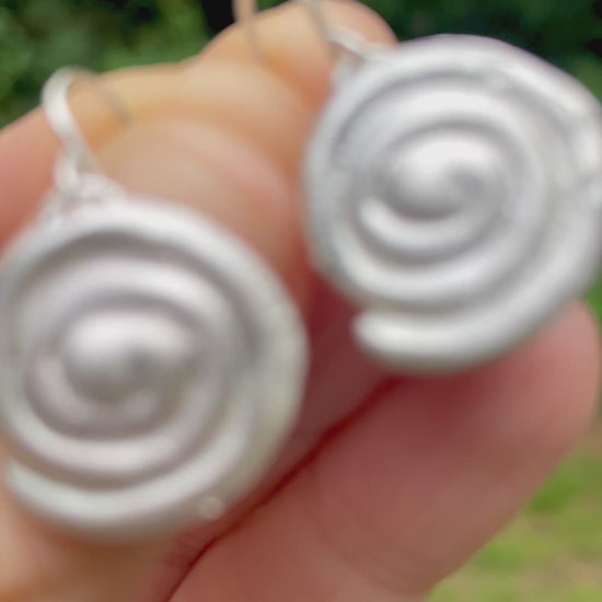 silver sandcast spiral earrings held in hand with green forest background