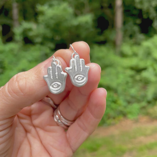 silver sandcast hamsa hand earrings held in a hand with green forest background