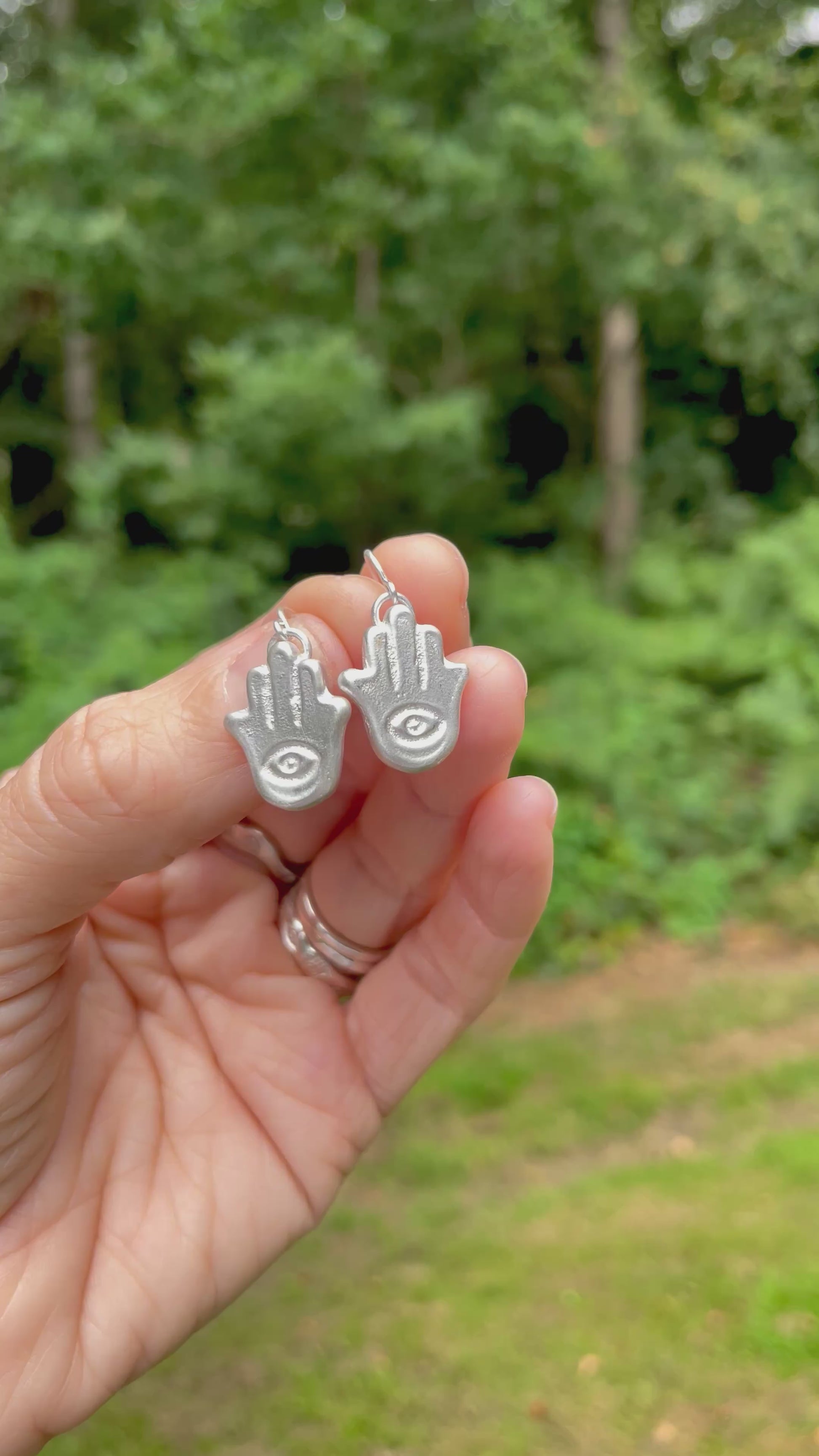silver sandcast hamsa hand earrings held in a hand with green forest background