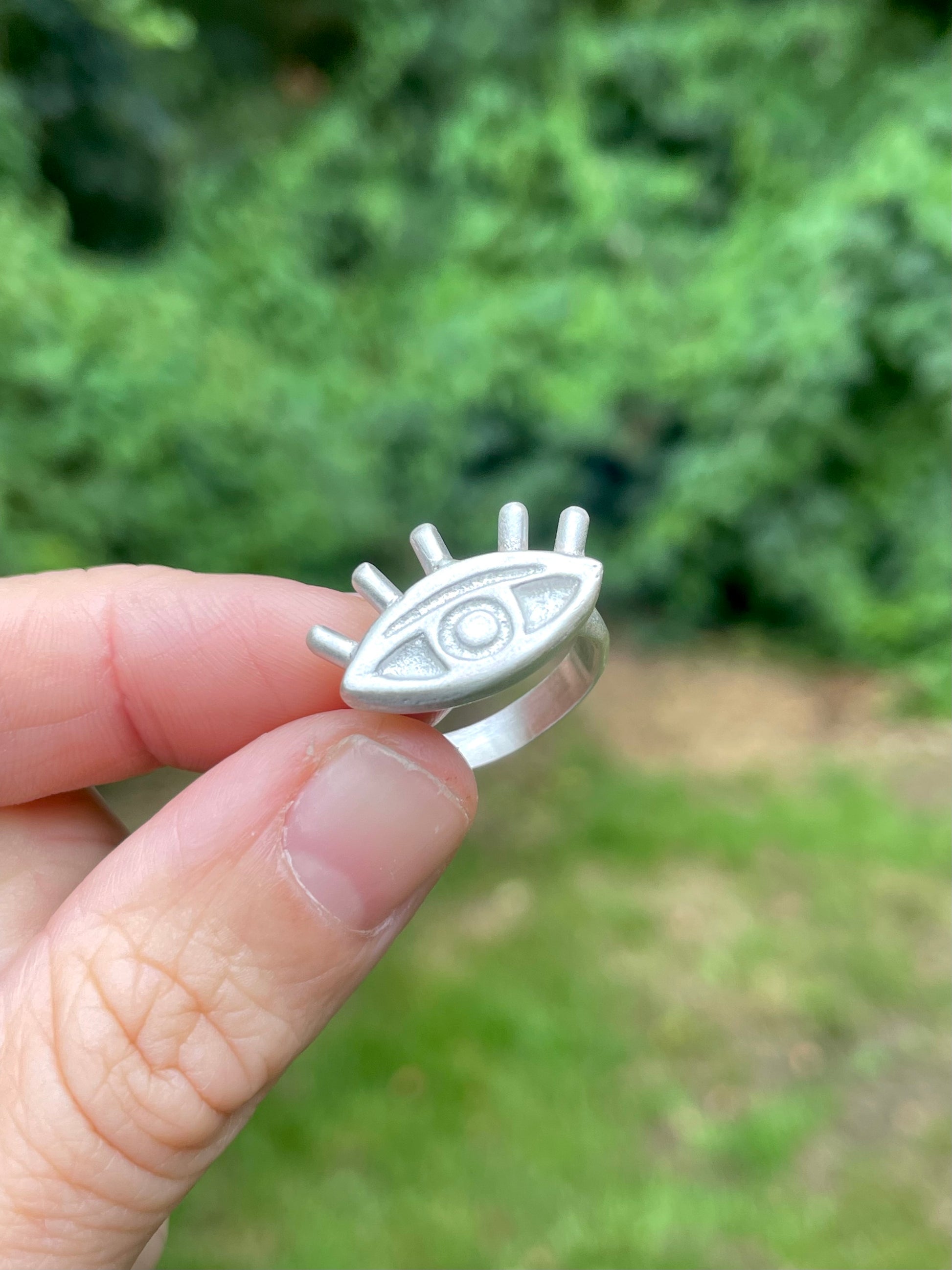 Silver sandcast ring with eye design held by a hand against a blurred green outdoor background