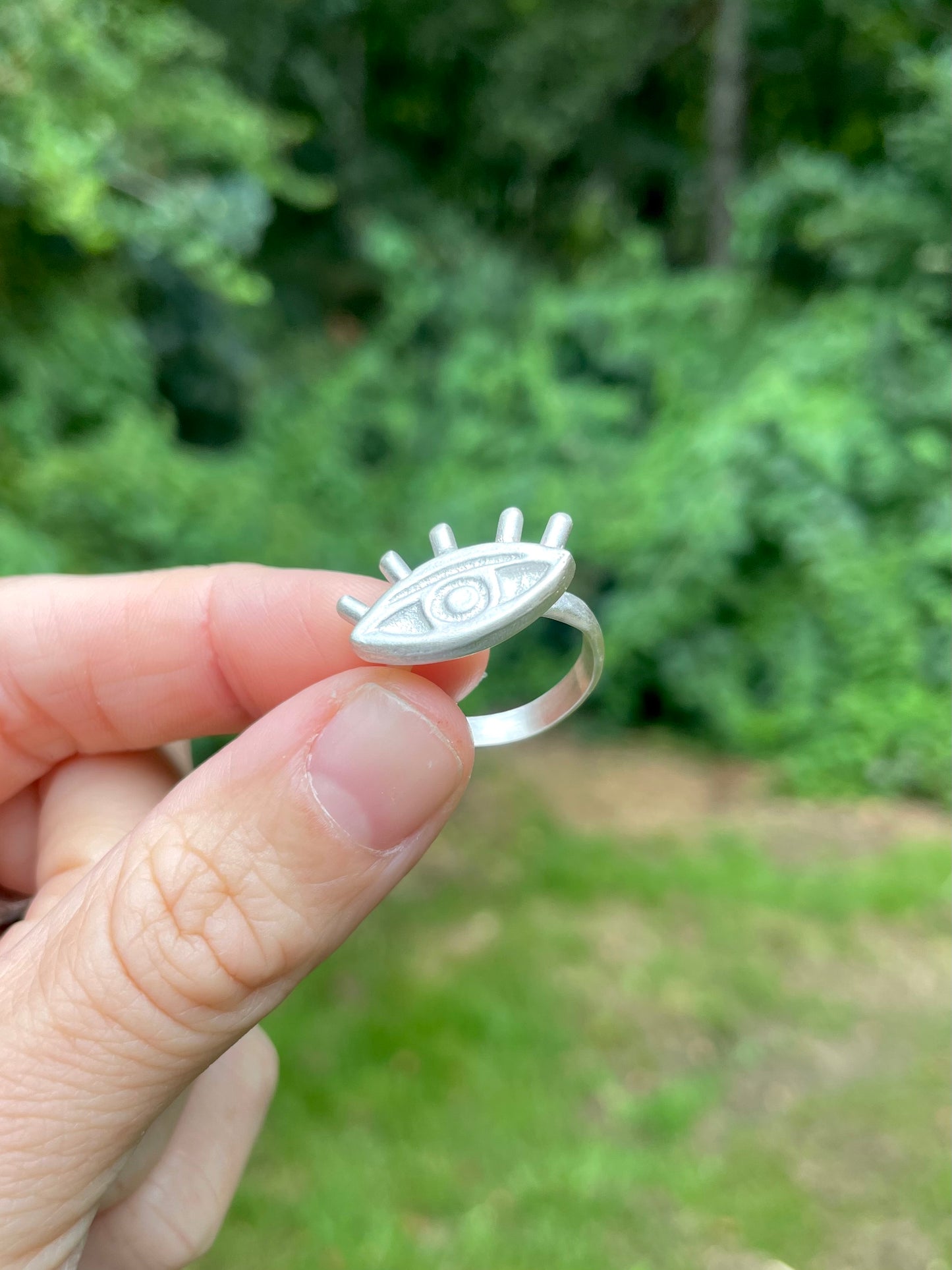 Silver eye ring held by a hand with a blurred green outdoor background