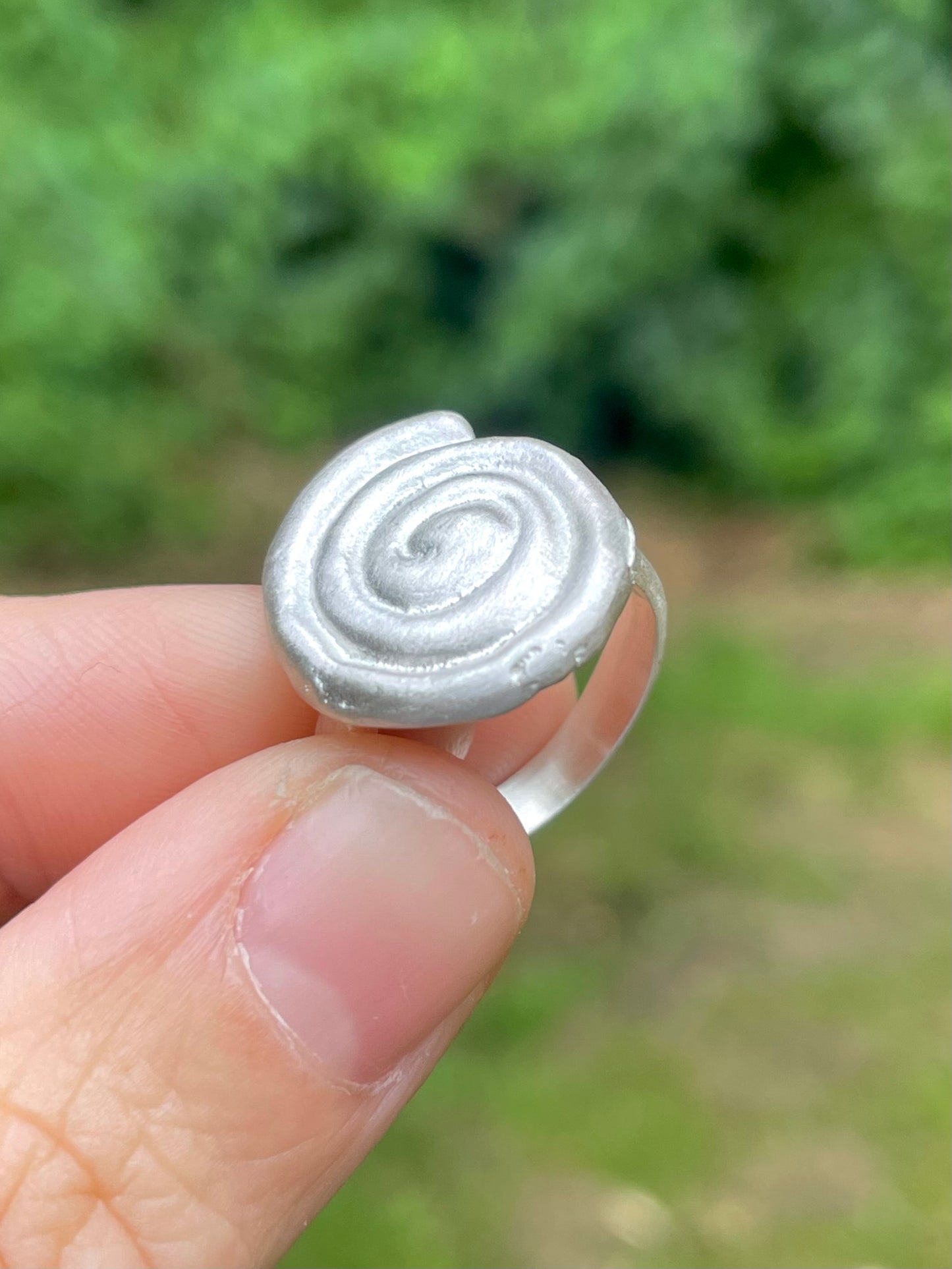Silver sandcast spiral ring held between fingers with a blurred green background
