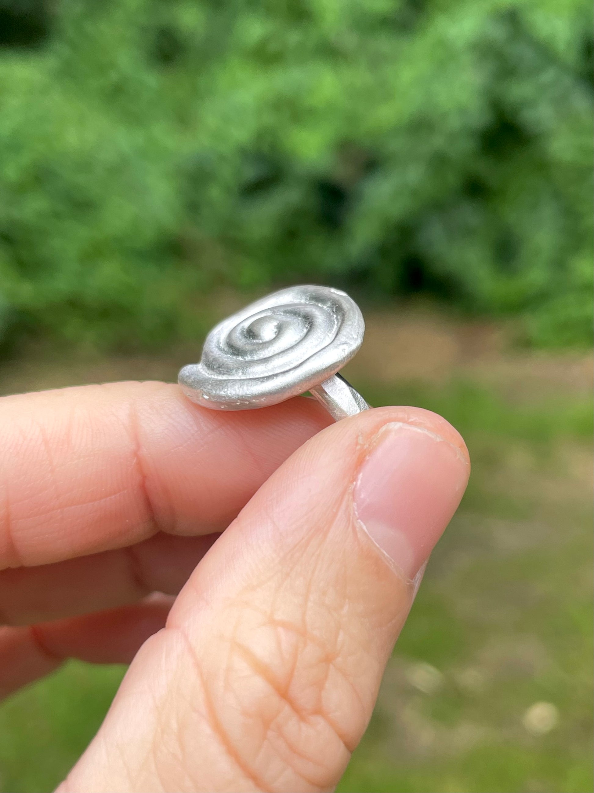 Silver sandcast ring with a spiral design held between fingers against a blurred green background