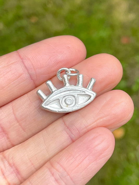 Silver eye-shaped pendant charm held between fingers with a blurred green background