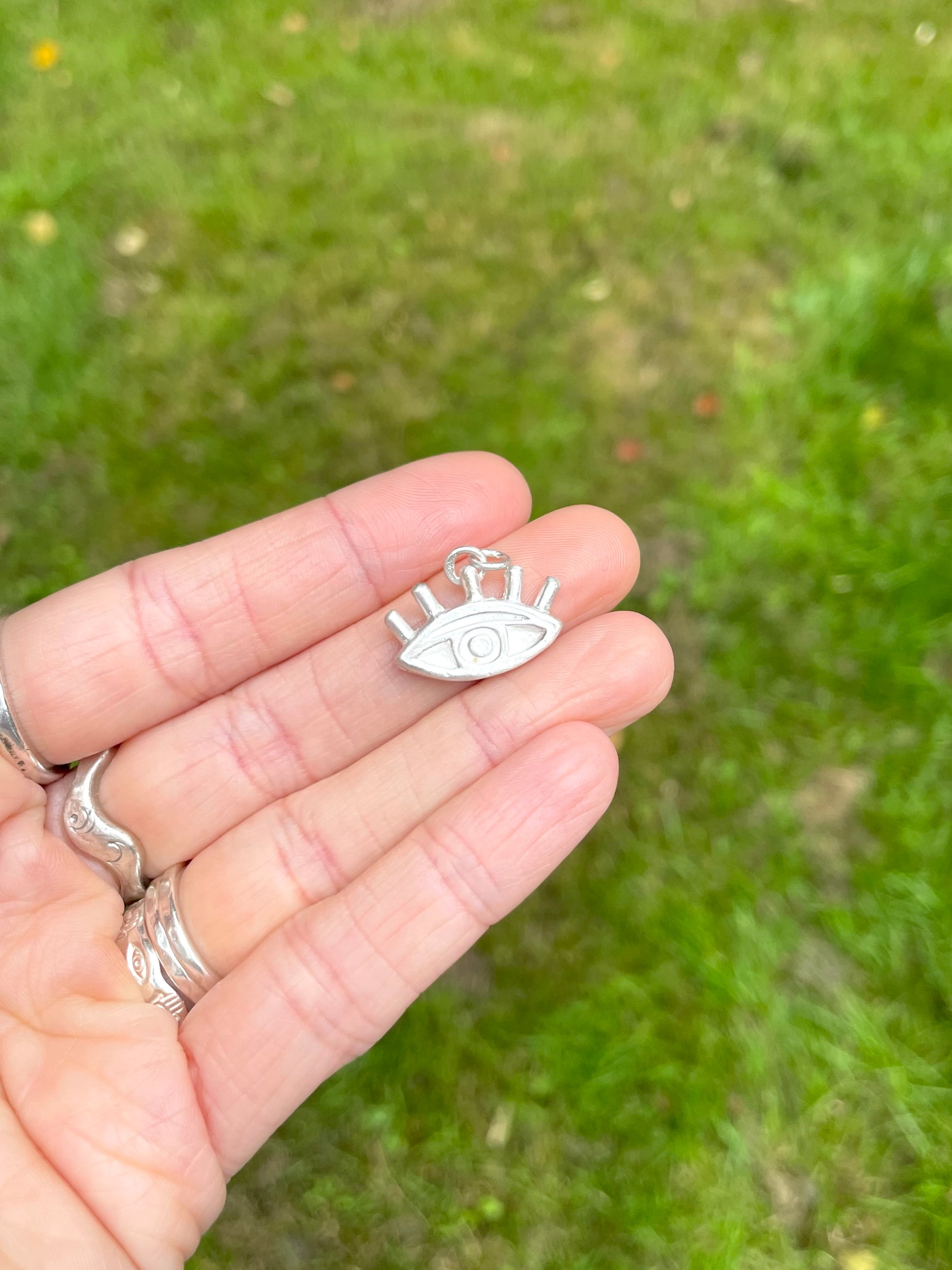 Small silver sandcast eye pendant held in a hand with a blurred green background