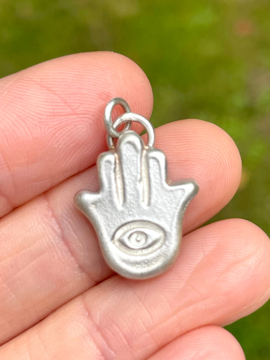 Silver sandcast Hamsa hand charm held between fingers with a blurred green background