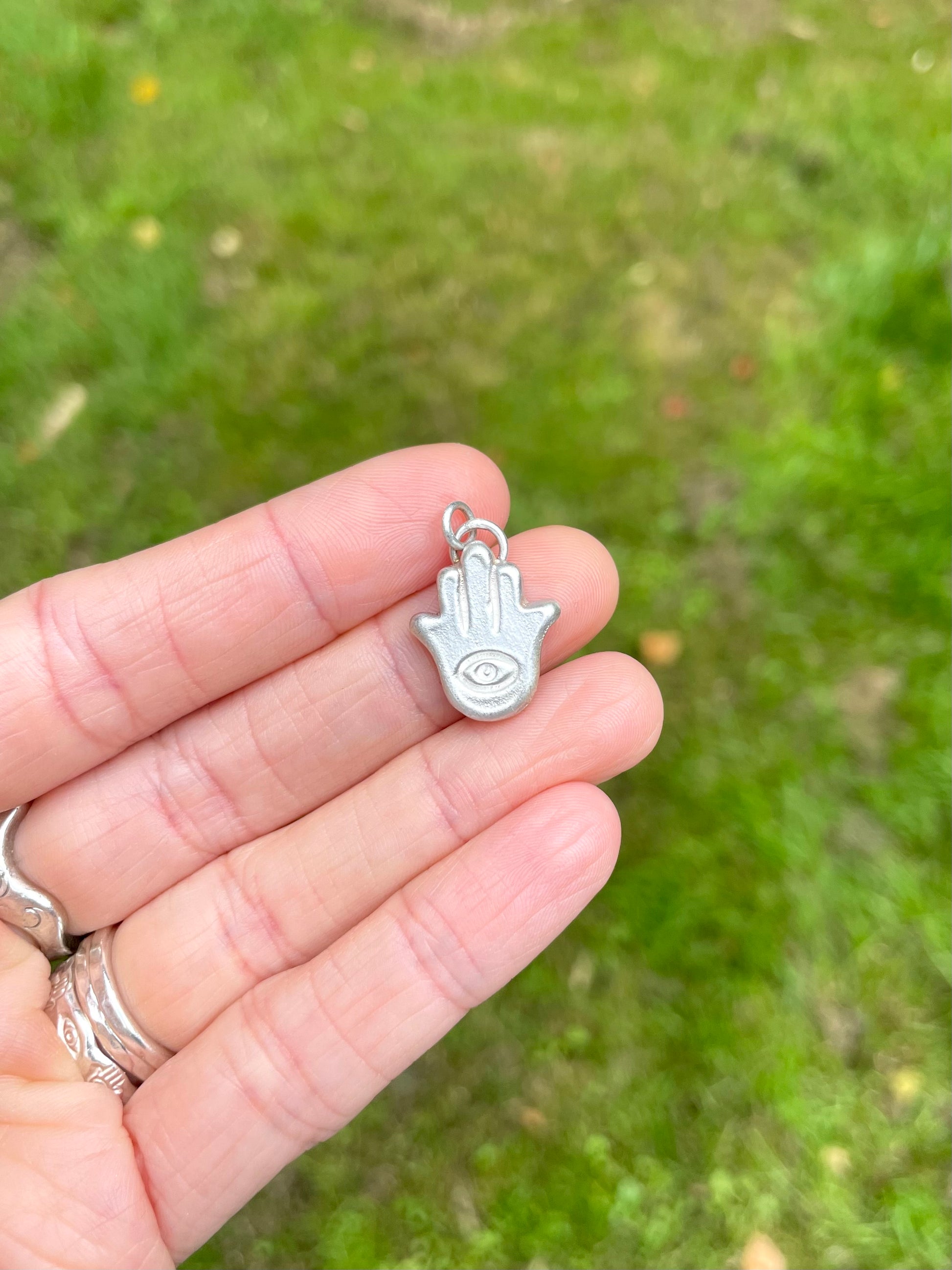 Silver sandcast Hamsa hand charm held in a hand against a grassy background