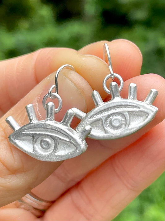 Silver sandcast eye-shaped earrings held in a hand with a blurred green background