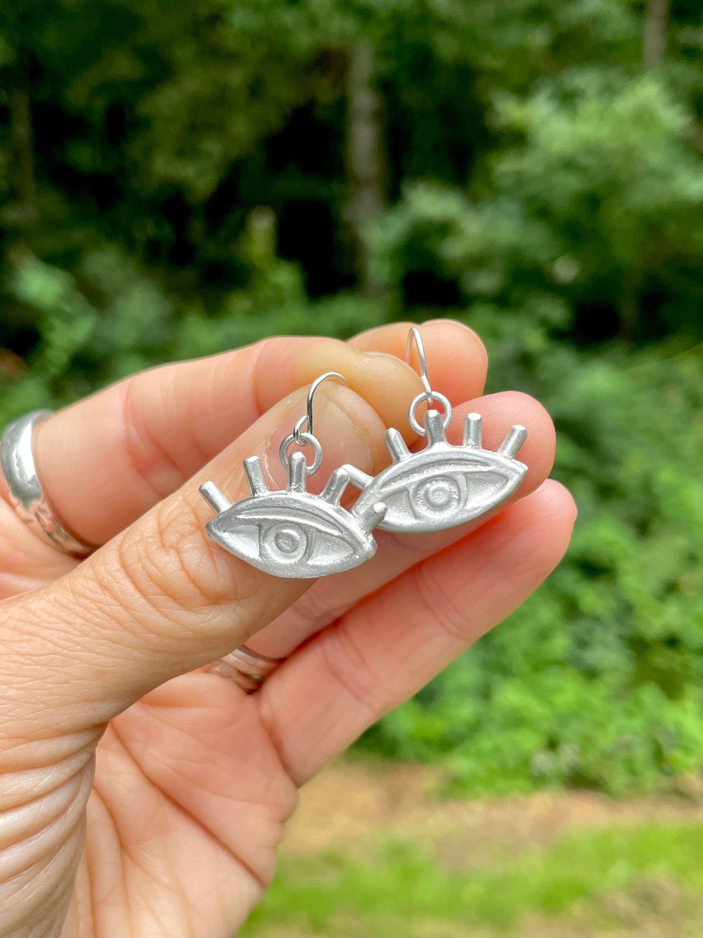 Silver sandcast eye-shaped earrings held in a hand with a blurred green outdoor background