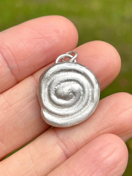 Silver sandcast spiral pendant held between fingers with a blurred green background