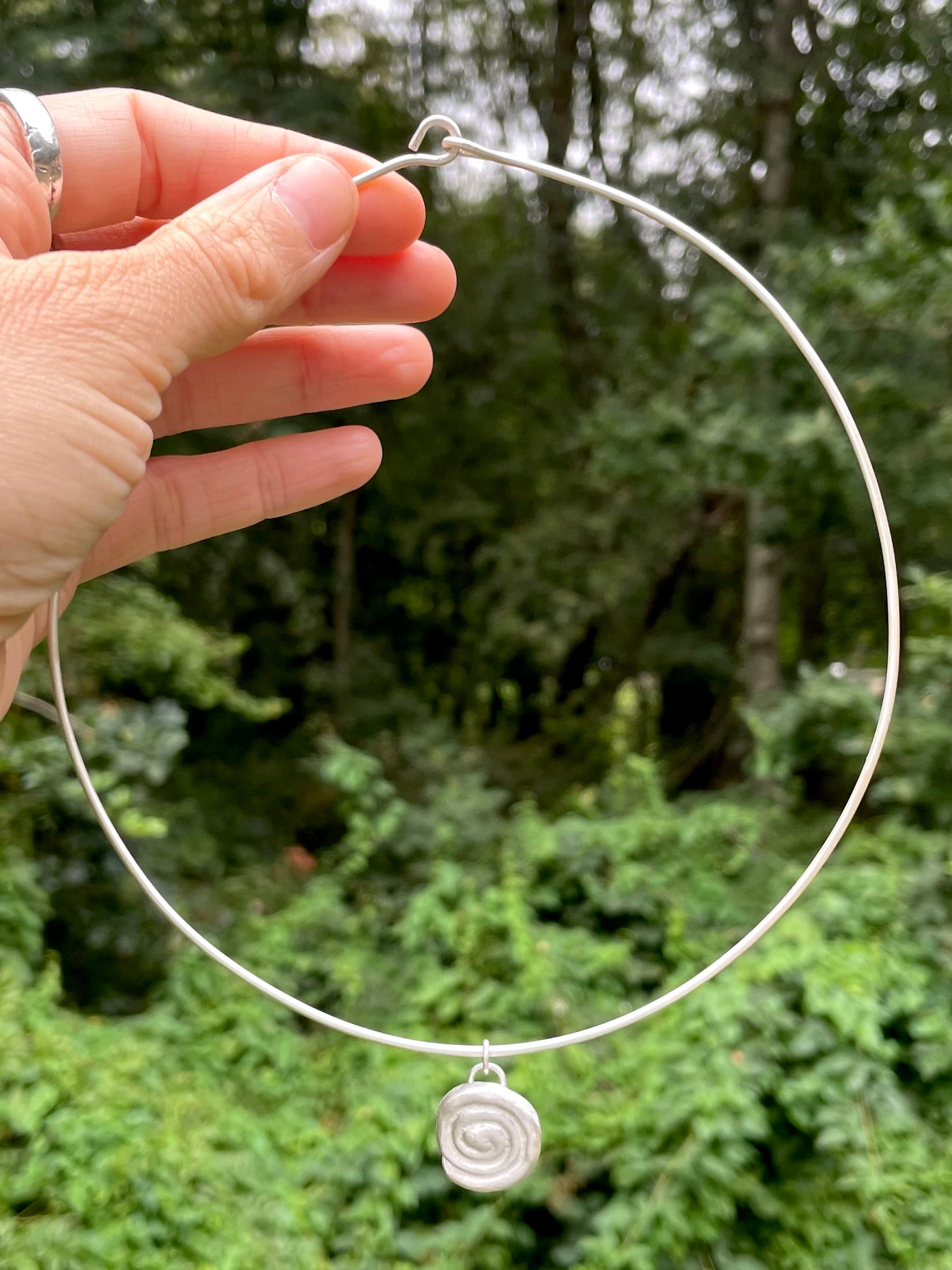 Silver choker necklace with a spiral sandcast pendant held by a hand against a green forest background